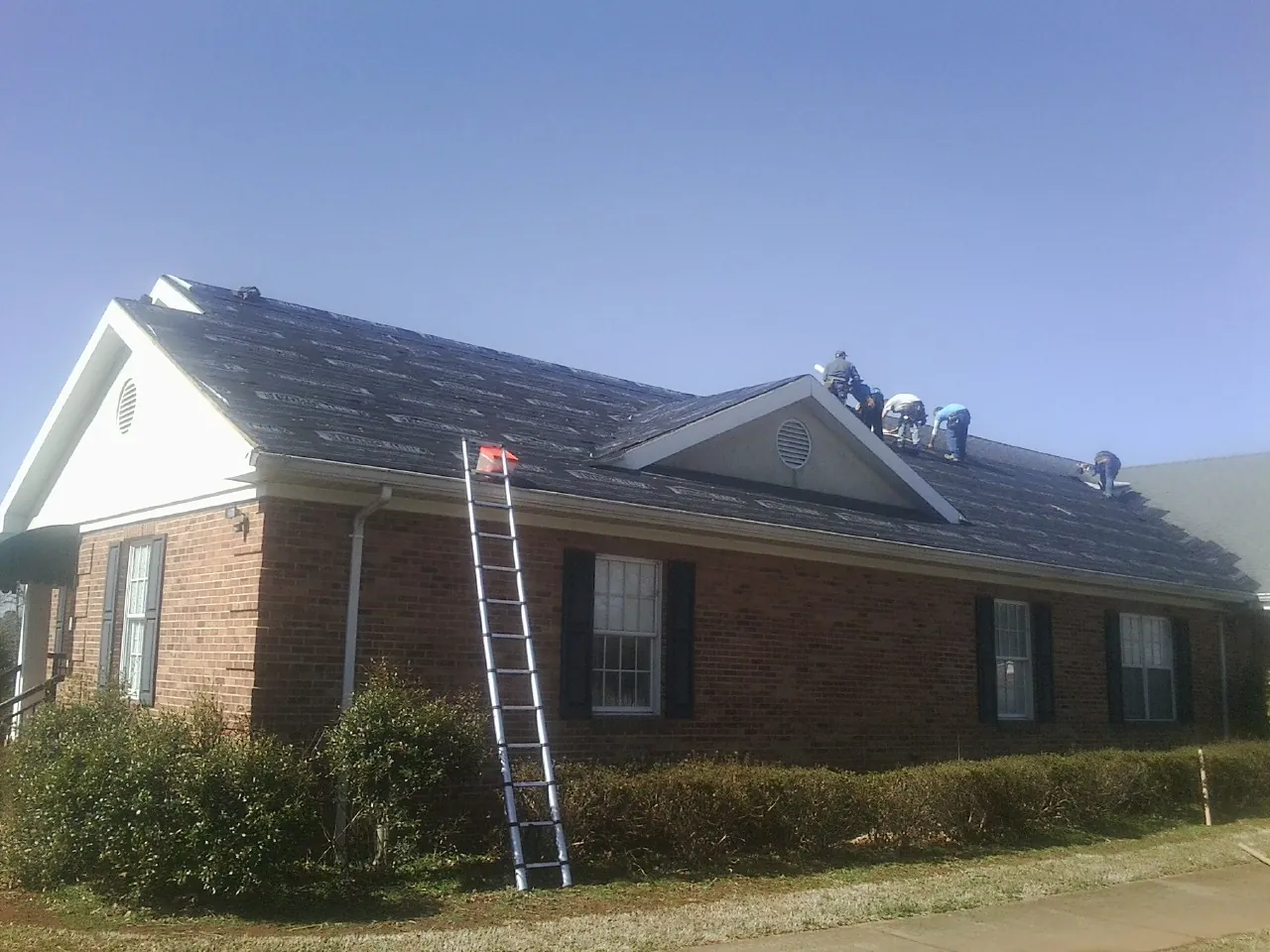 Crew tearing shingles off a brick ranch roof with a ladder set — Metal Roof Repair work by our crew in Revloc
