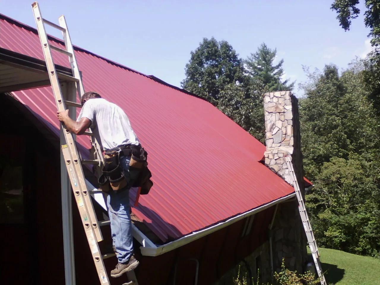 Roofer on a ladder beside a red metal roof and stone chimney — Soffit & Fascia Repair by Roofing Brothers in Revloc